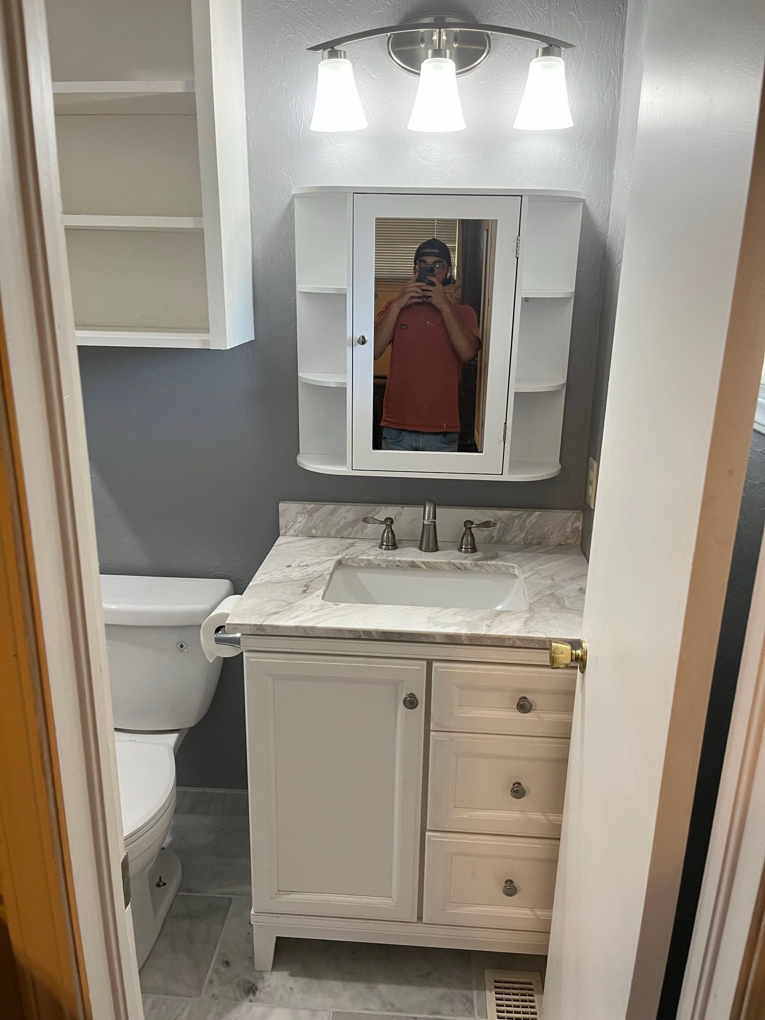 Small bathroom with a white vanity, toilet, and medicine cabinet; a person is reflected in the mirror.