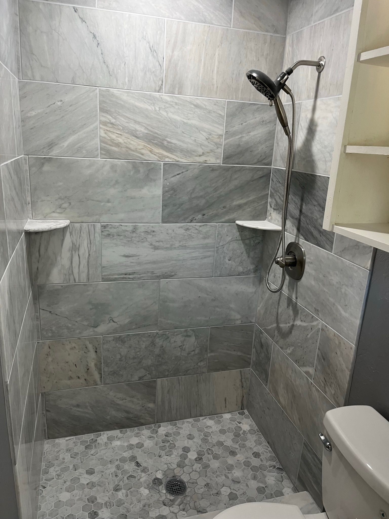 Gray-tiled shower with built-in shelves, pebble floor, and showerhead. Toilet is partially visible on the right.