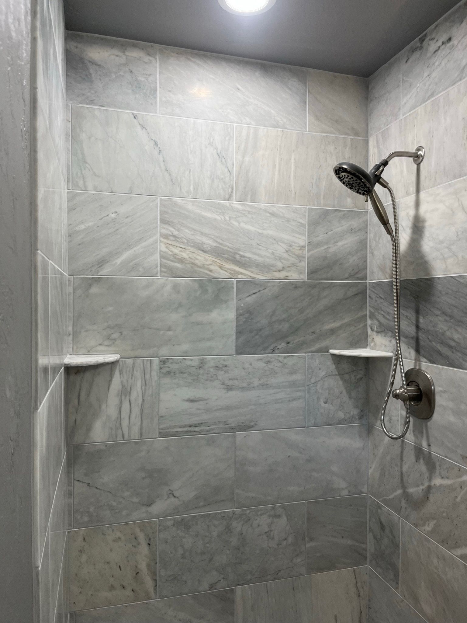 Gray and white tiled shower with built-in shelves, a showerhead, and a handheld shower.