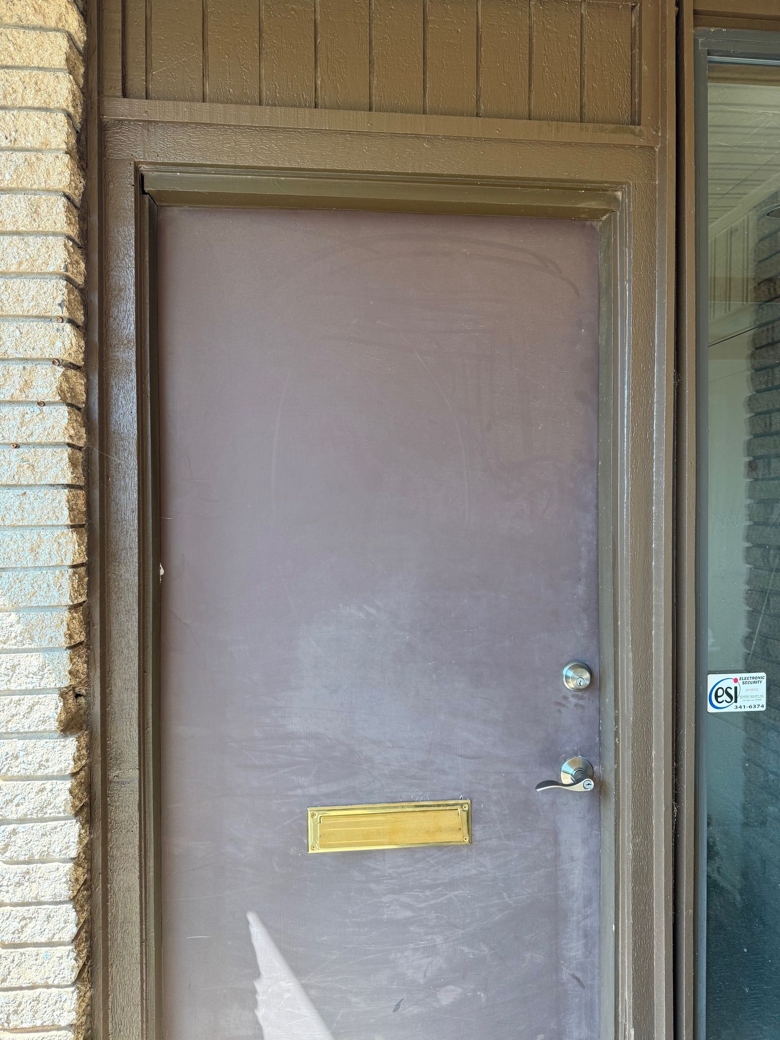 Brown door with gold mail slot, lock, and handle. Brick wall on the left, glass on the right.