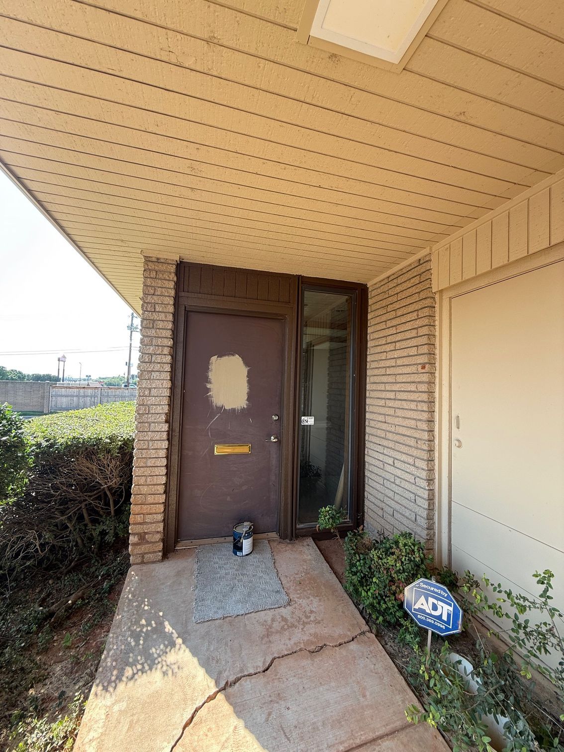 Brown front door with cracked concrete pathway, covered by a tan awning, and overgrown foliage.