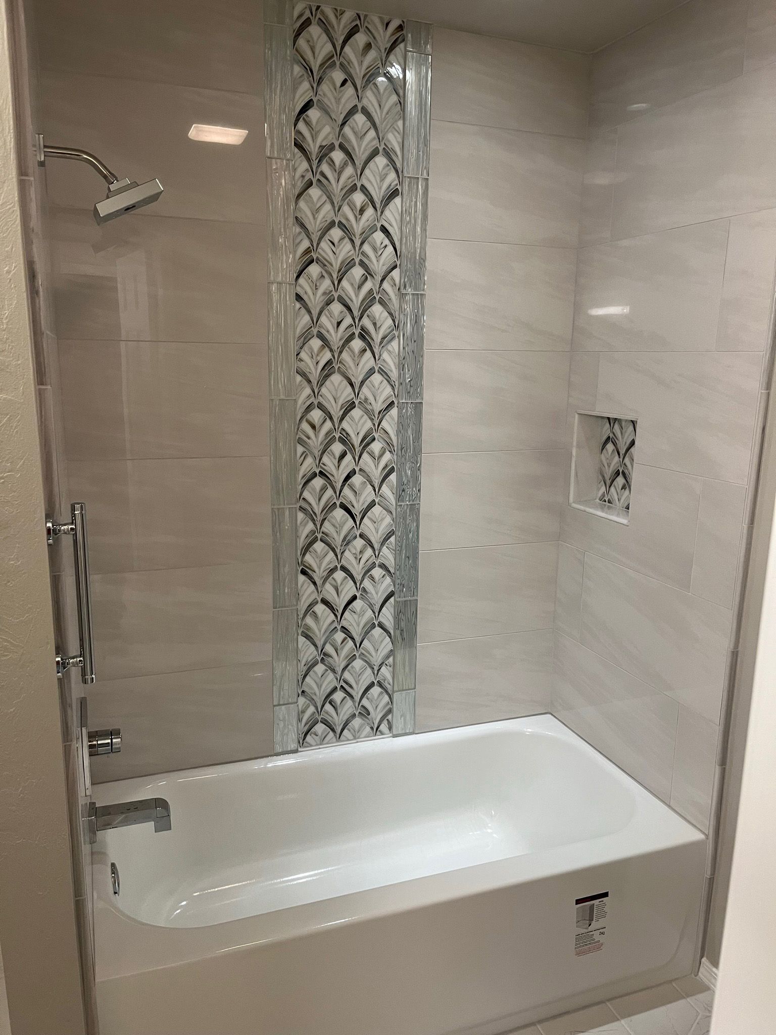 A white bathtub and shower with a decorative tile strip and a built-in niche.