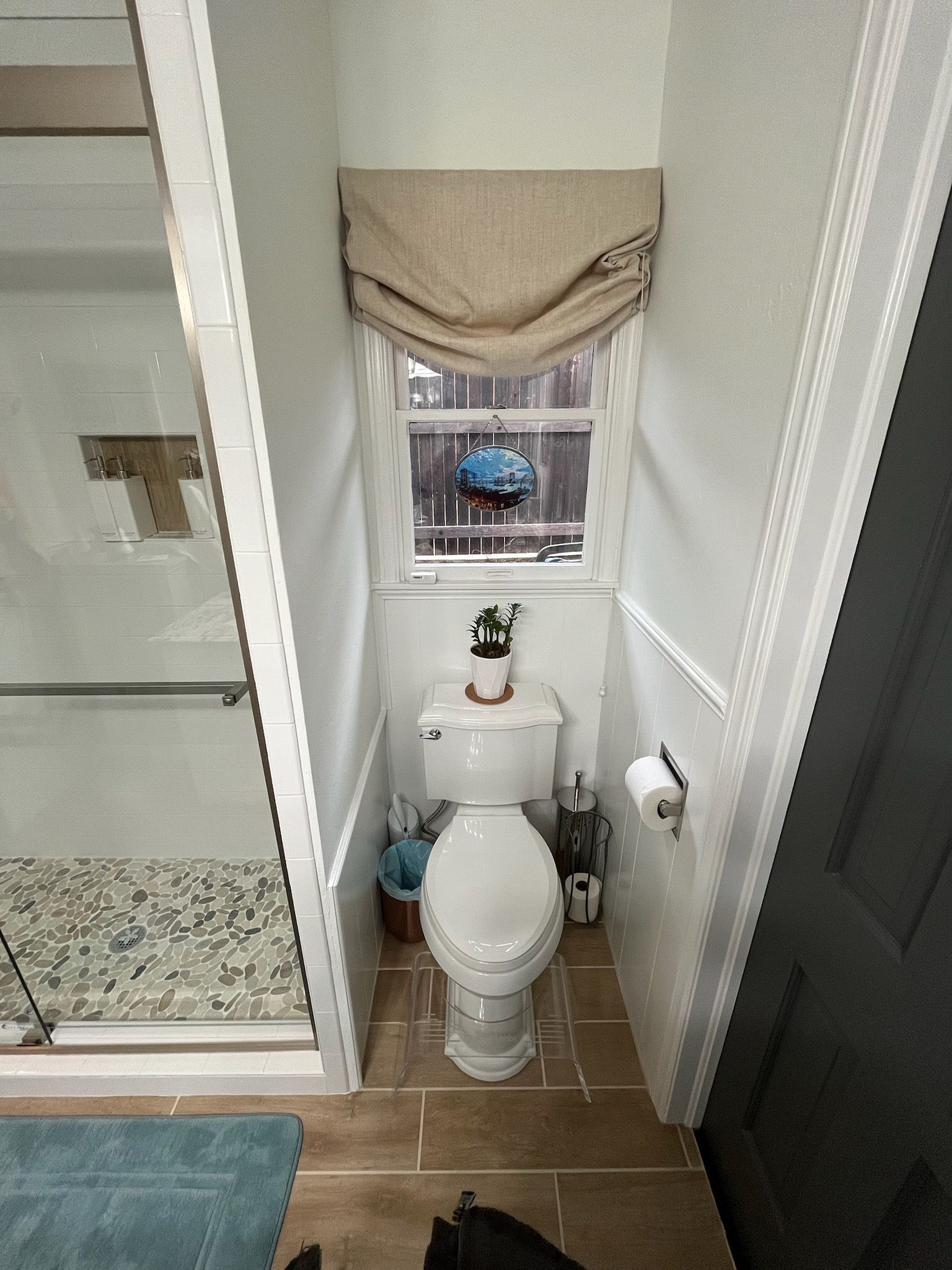 Small bathroom with toilet under a window. Beige roman shade, white walls, and wood-look tile.
