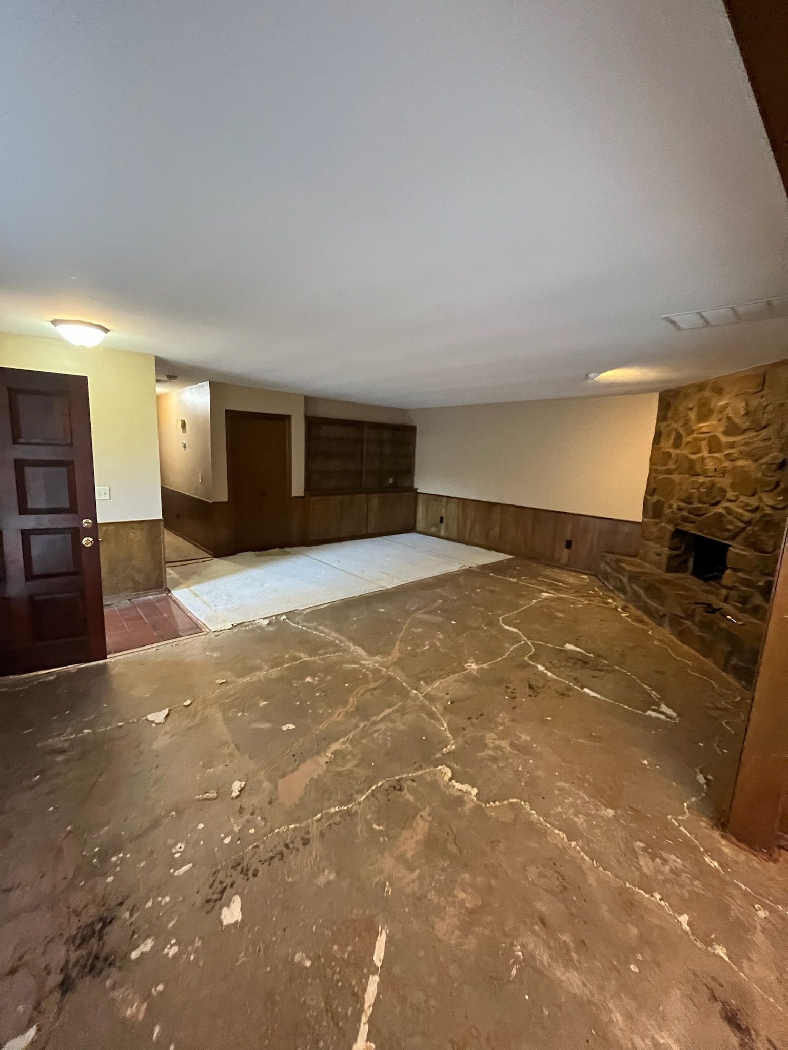 Empty room with damaged concrete floor, stone fireplace, and wood-paneled walls.