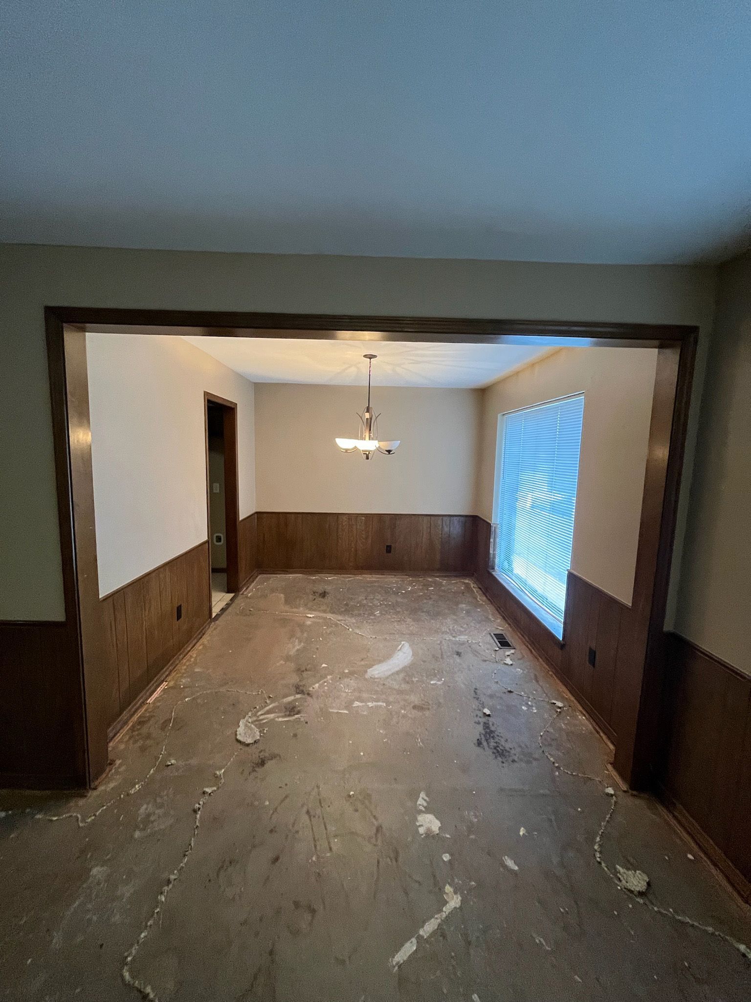 Empty room with dirty floor, wood paneling, and an open doorway. Natural light streams through a window.