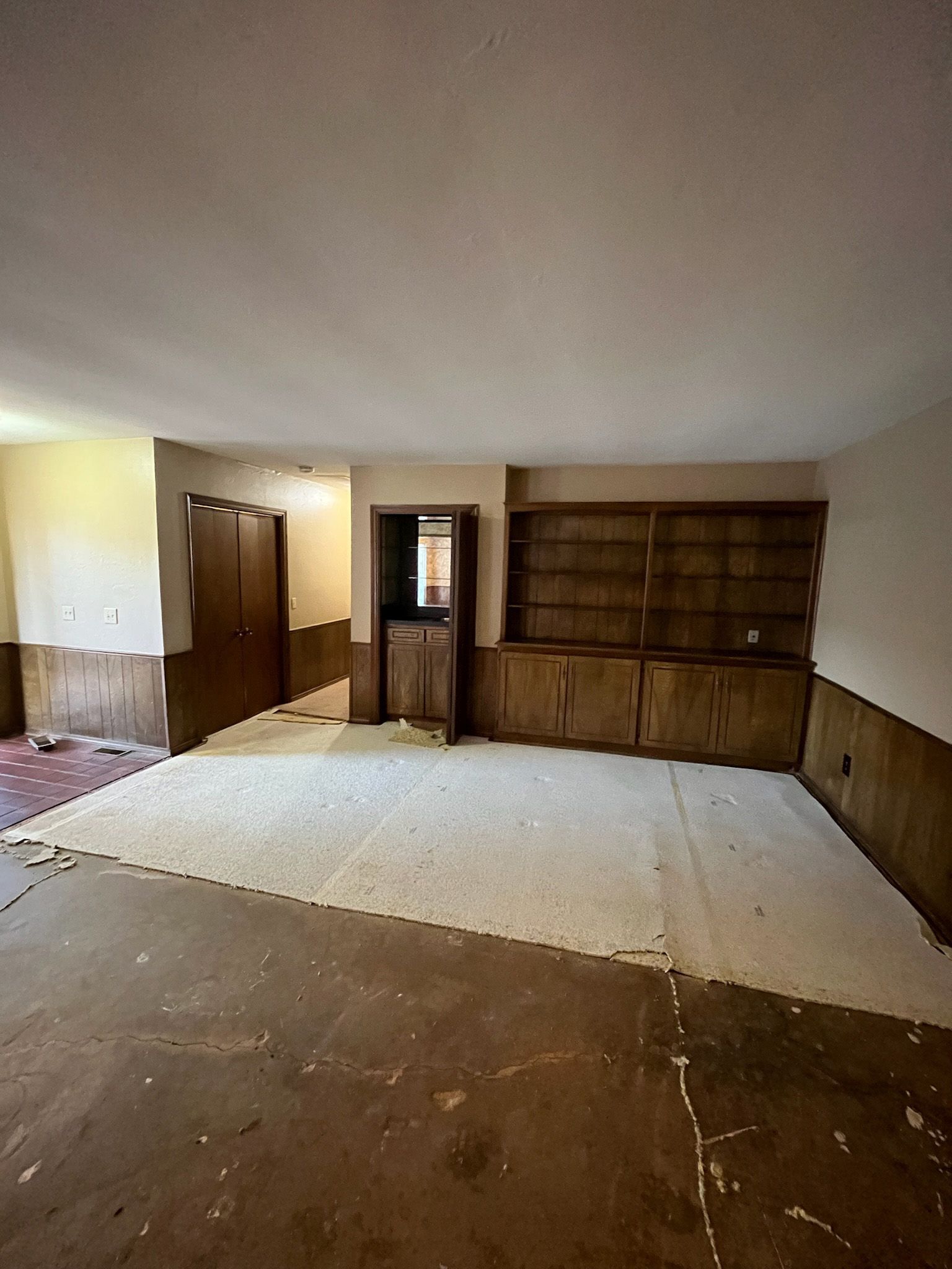 Room with damaged flooring, built-in shelves, and a cabinet. Wood paneling on lower walls.