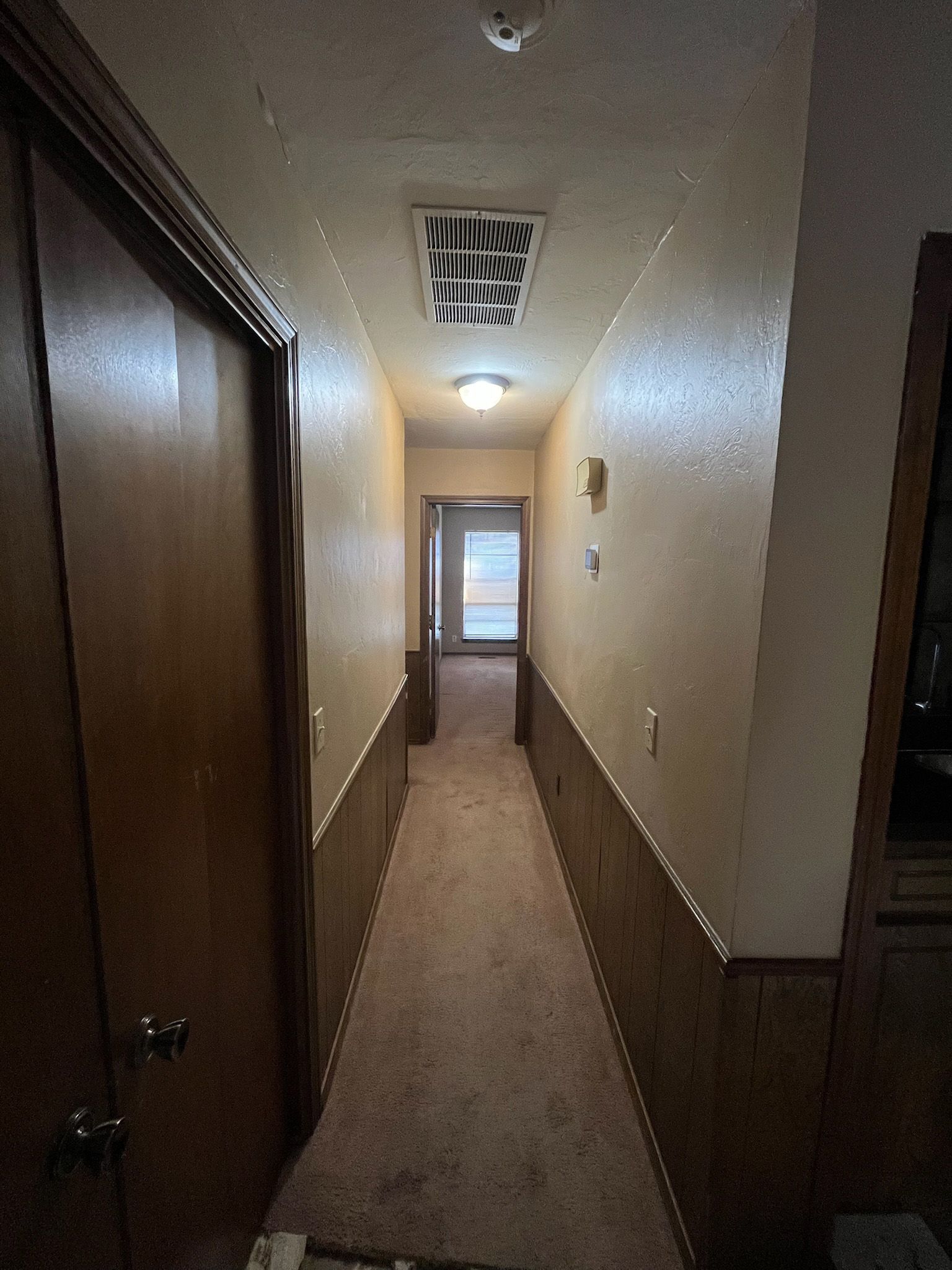 Narrow hallway with wood doors, paneling, and carpet; a light fixture on the ceiling.