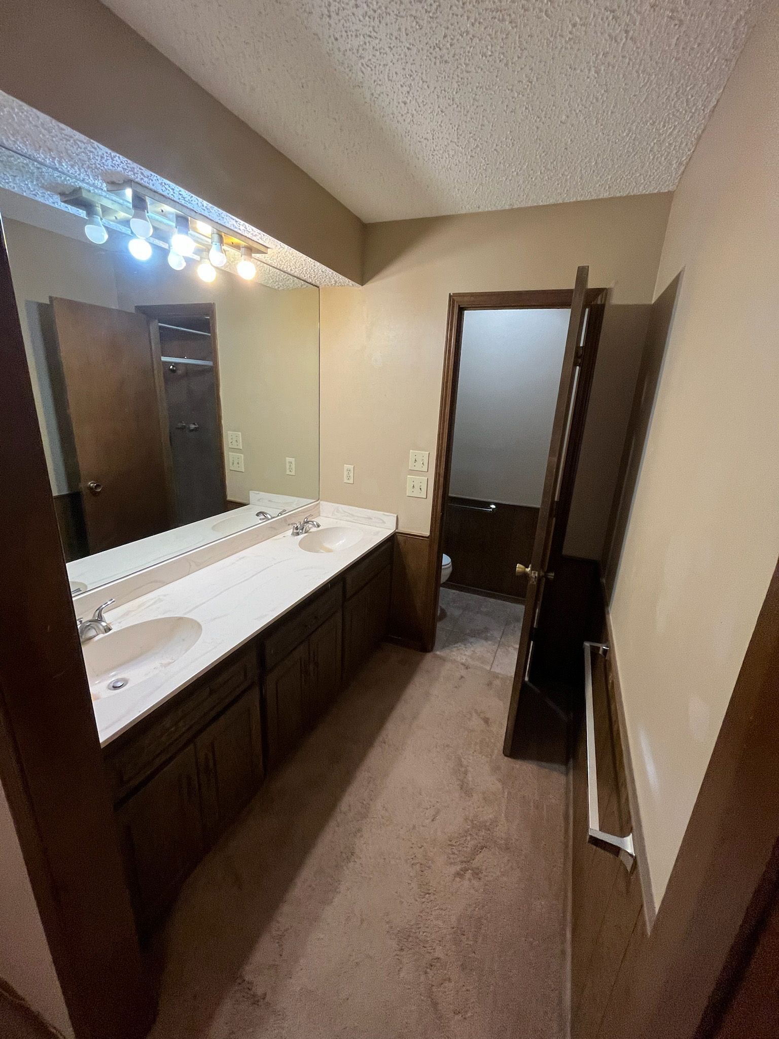 Bathroom with double vanity, brown cabinets, doorway to a toilet, and a long, narrow hallway.