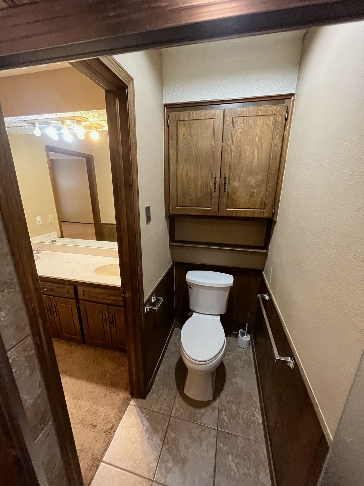 A small bathroom with a toilet, storage cabinet, and doorway to a vanity area with brown trim.