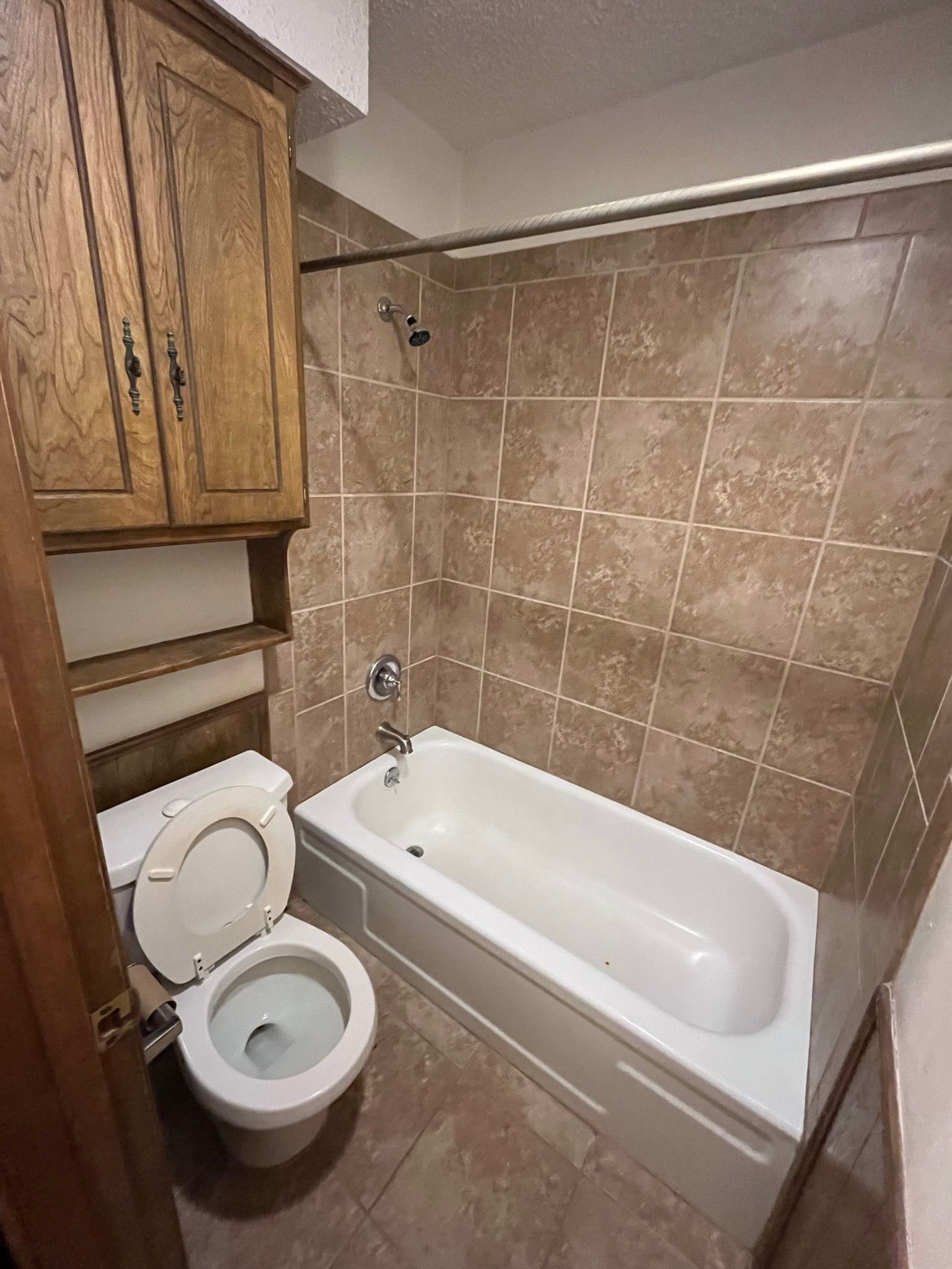 Bathroom with toilet, bathtub, brown tile, and wooden cabinet.