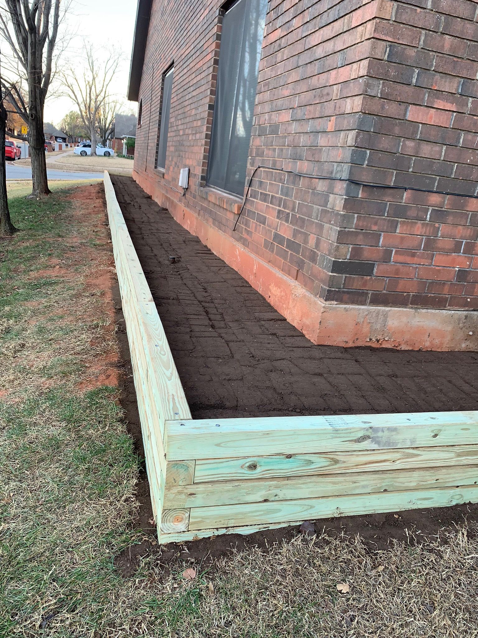 A raised garden bed constructed of green-treated wood against a brick building, filled with dark soil.