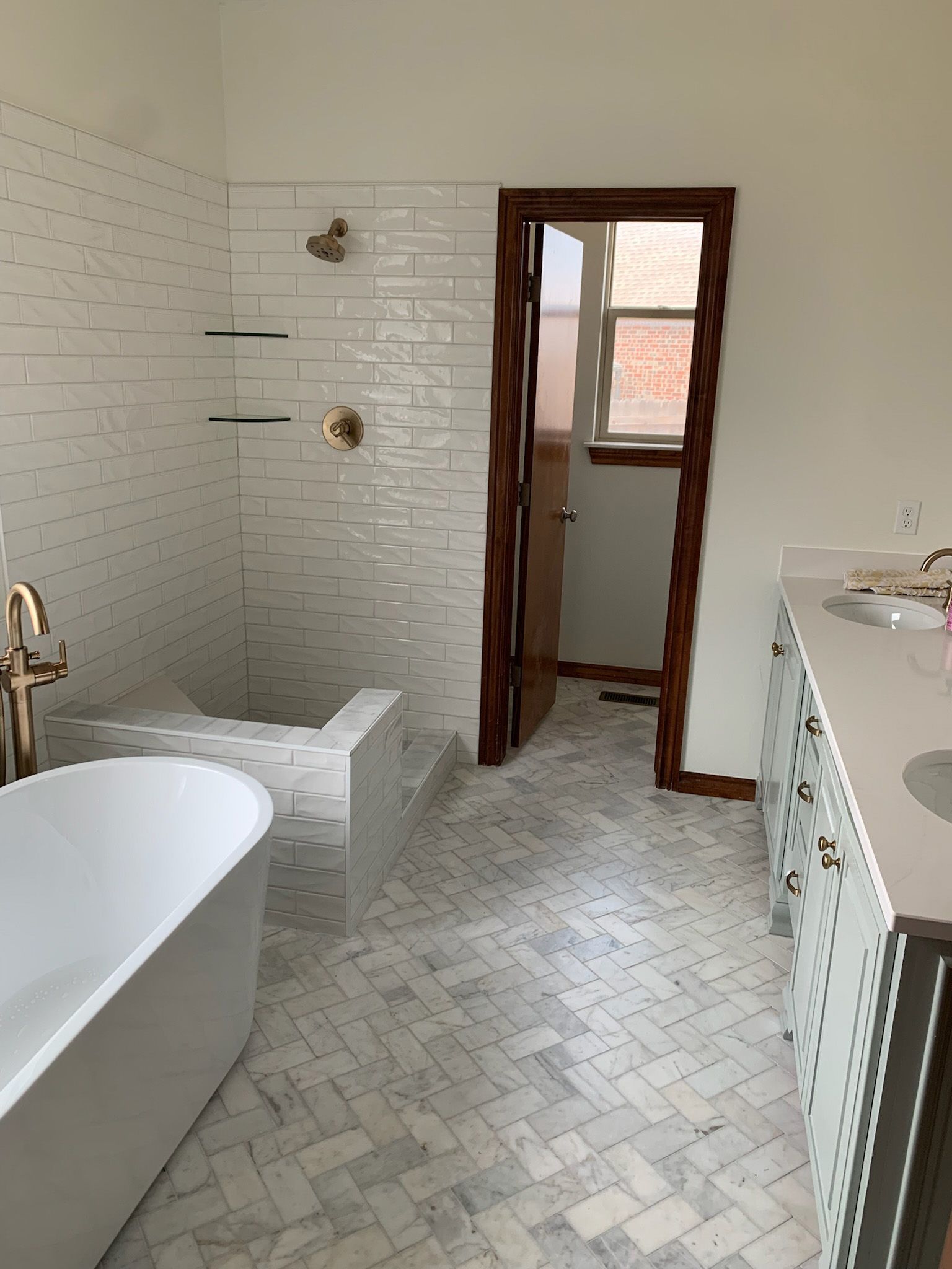 White bathroom with herringbone tile floor, freestanding tub, and wooden door.