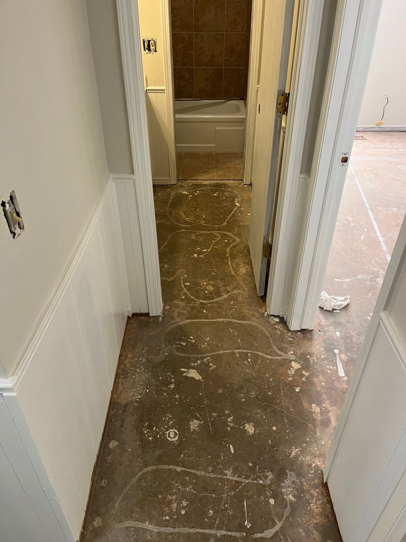 Narrow hallway with damaged flooring, leading to a bathroom with a tub. White walls and door frames.