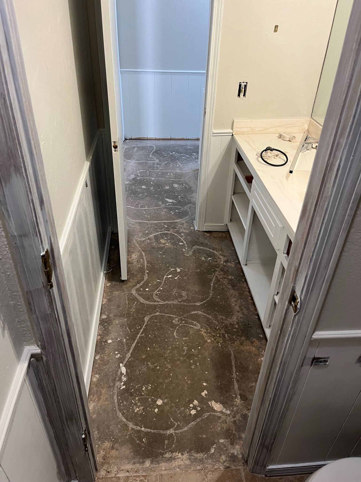 Bathroom with open doorway, floor with old glue residue, white cabinet and mirror, and doorway to another room.