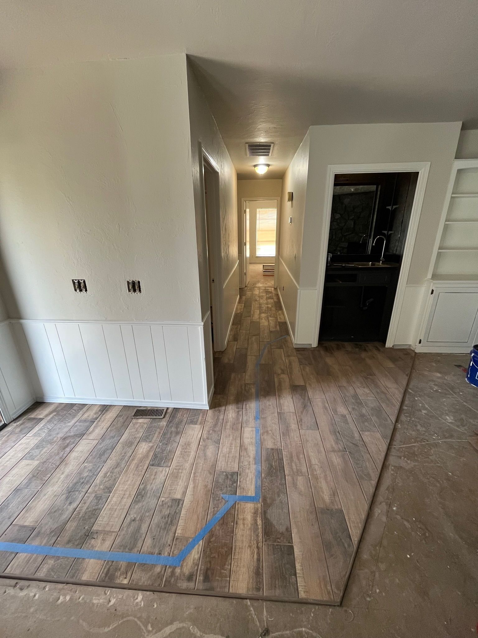 A partially renovated interior shows wood flooring, a hallway, and a kitchen. Blue tape marks a floor area.