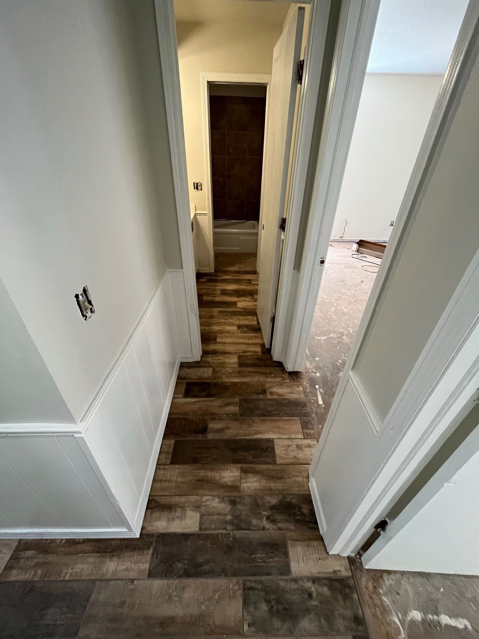Narrow hallway with wood-look flooring, white wainscoting, and open doors leading to a bathroom and a room.
