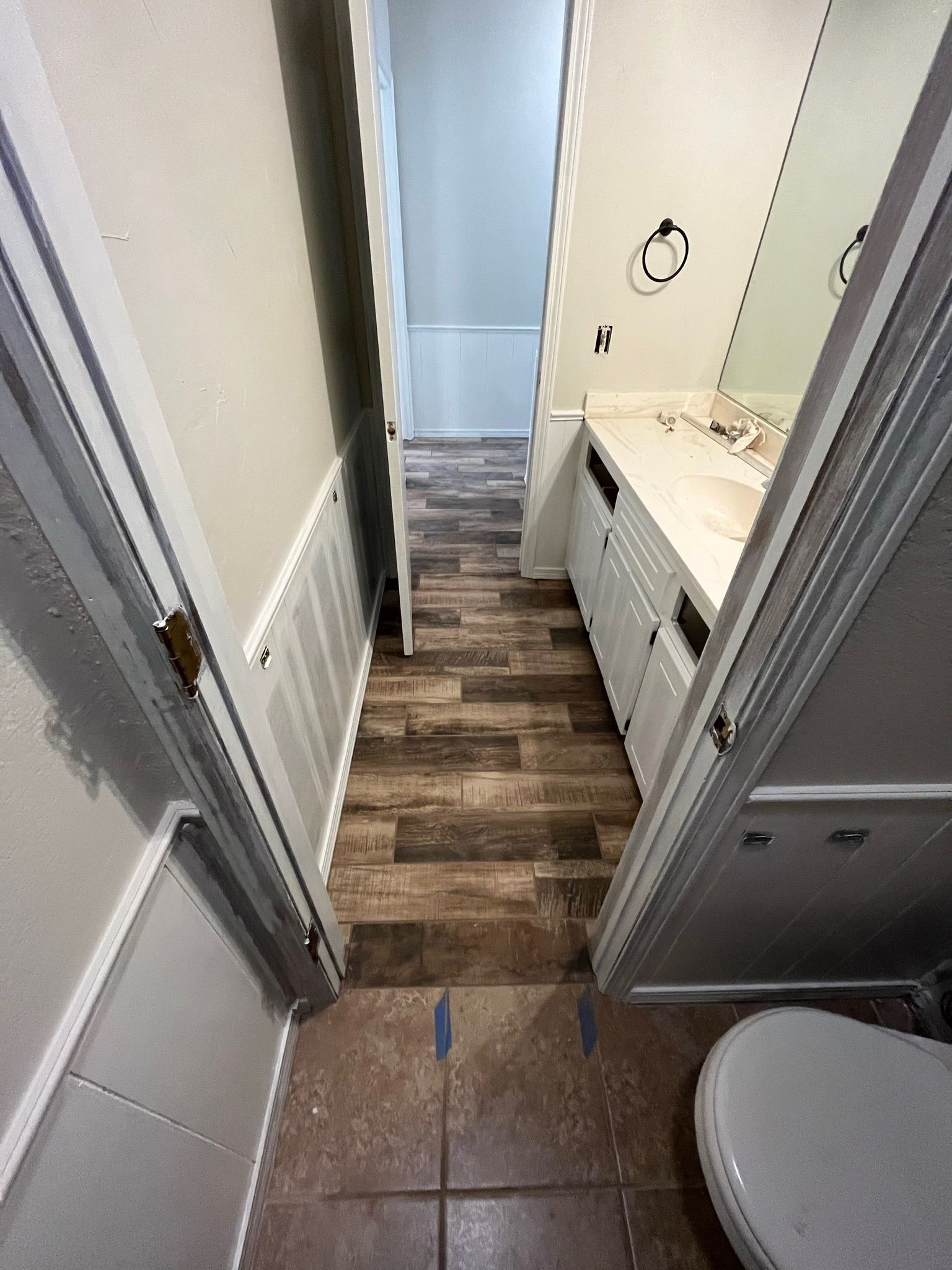 Narrow bathroom with a toilet, sink, and doorway to a room with wood-look flooring.