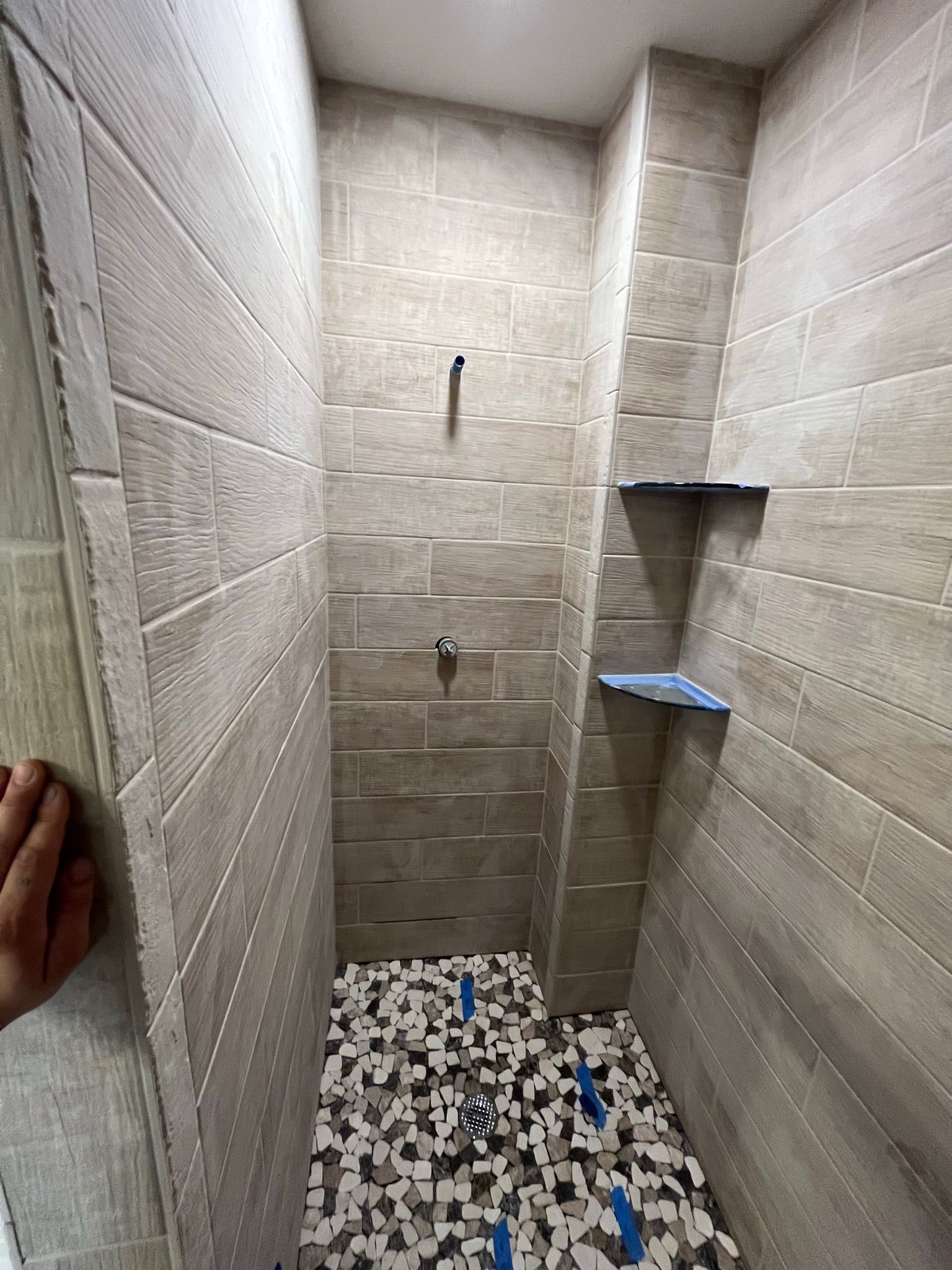 A narrow, tiled shower with neutral walls and a mosaic floor. Two shelves are visible.