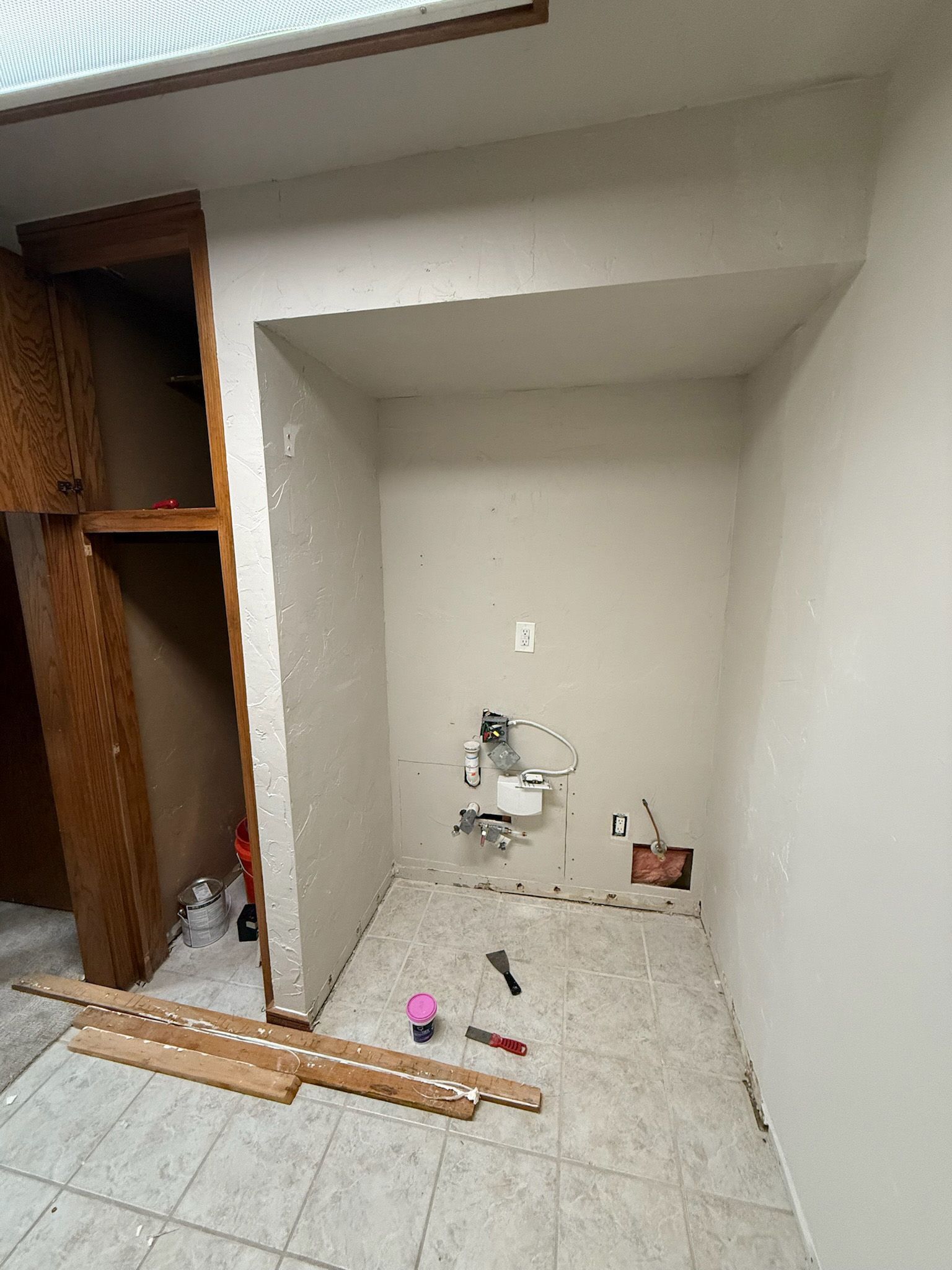 Laundry nook under construction, with exposed plumbing and floor tiles. Wooden cabinets and trim on the left.