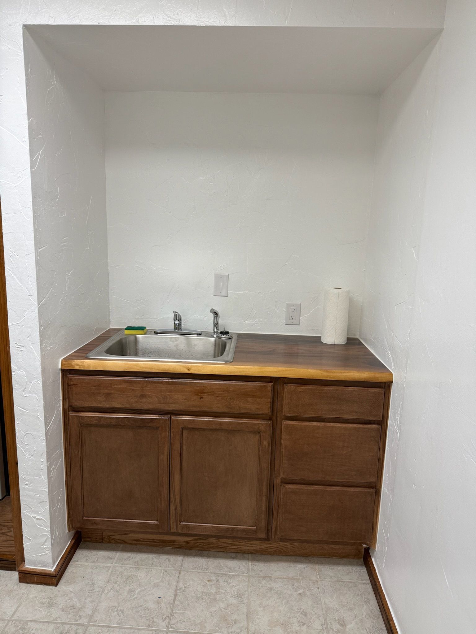 Brown cabinet with sink, paper towel, and outlet on white walls.