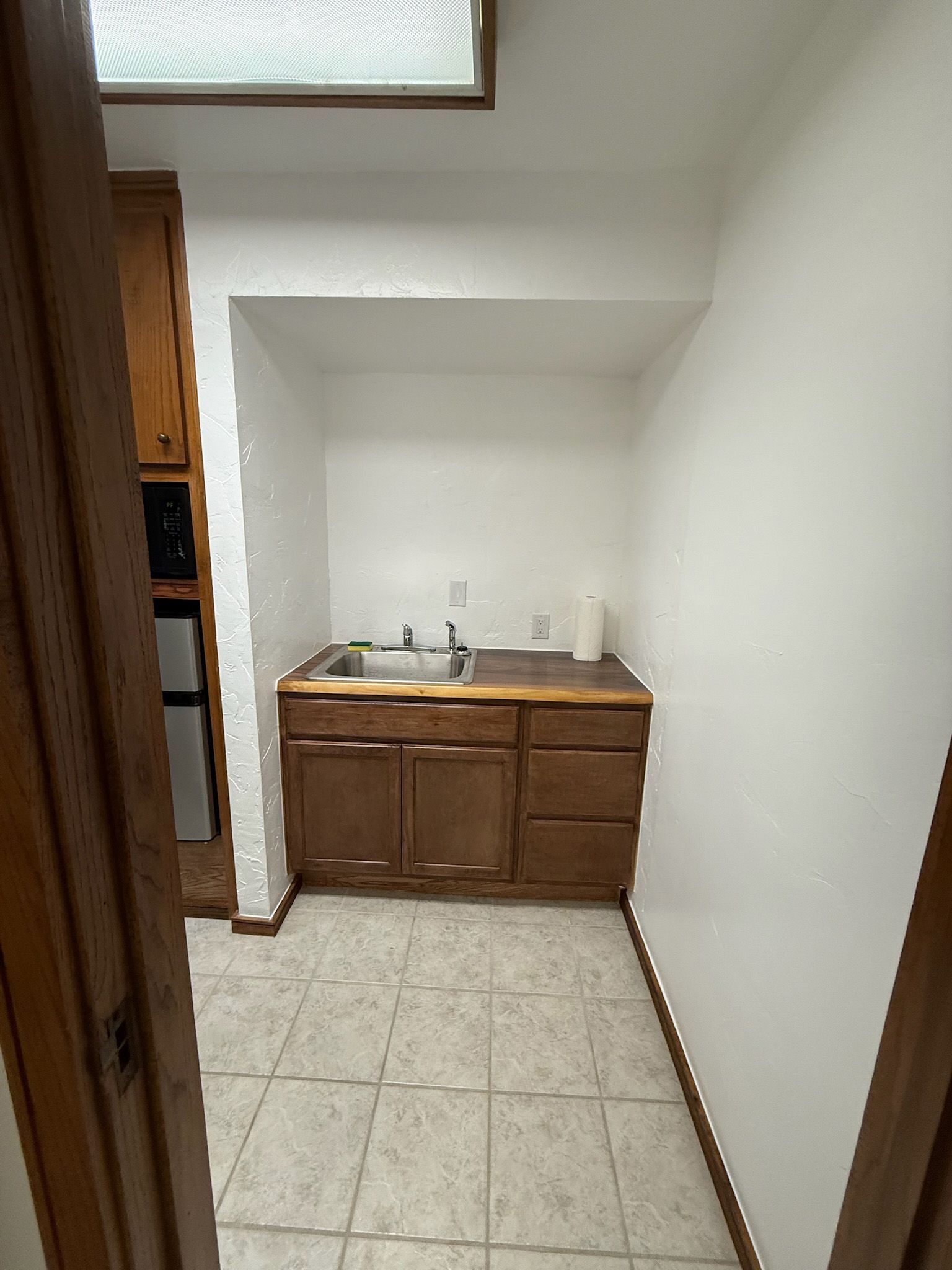 Small kitchenette with wooden cabinets, sink, and microwave; tile floor.