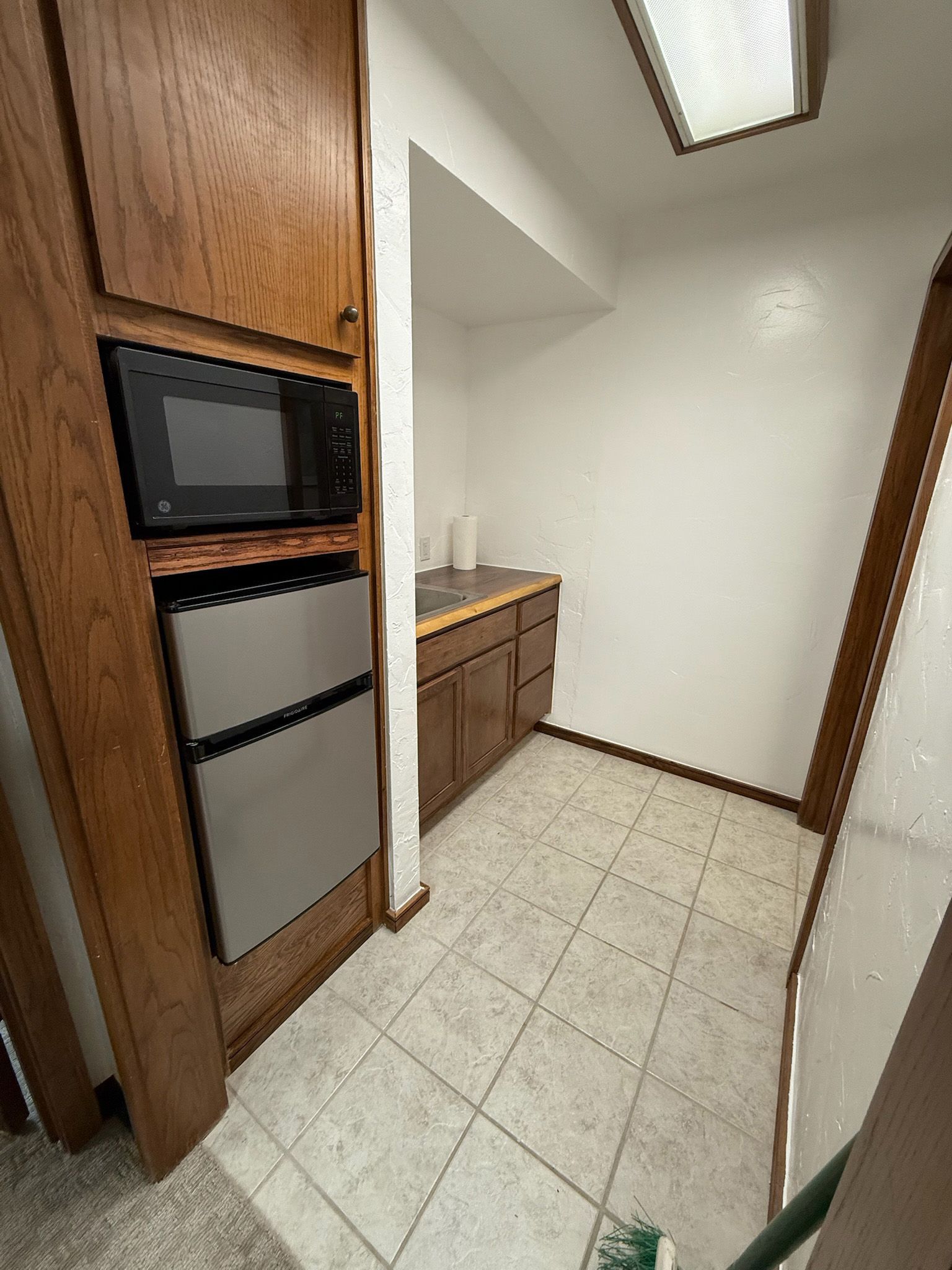 Small kitchenette with a microwave, mini-fridge, cabinets, and a sink. Light tile floor, white walls.