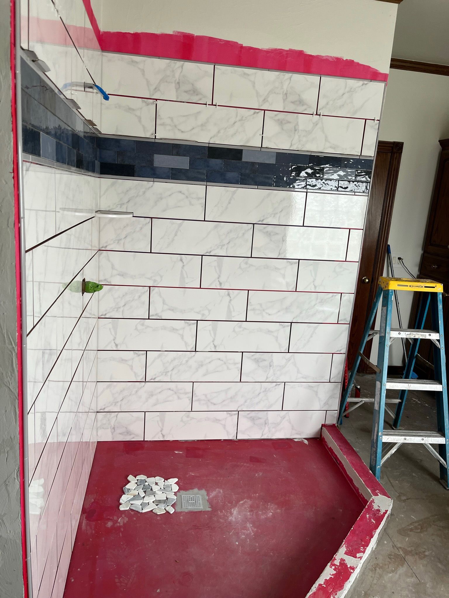 Shower stall under construction with white, dark blue, and pink tiles. A ladder stands nearby.