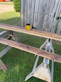 Wood planks stained brown on sawhorses outside. A can of stain and brush are on the planks near a wooden fence.