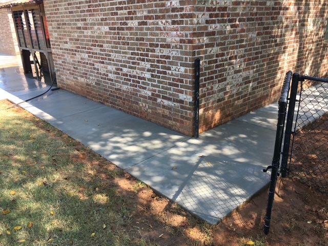 Concrete walkway beside a brick building, leading to a black fence. Sunlight casts shadows.
