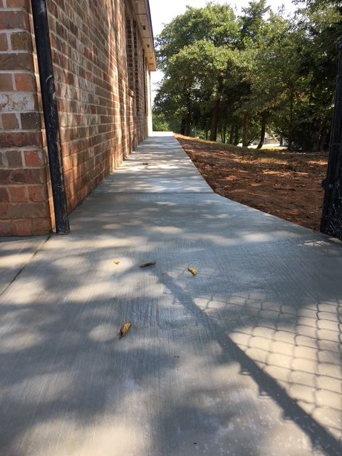 Concrete walkway next to a brick building and a chain-link fence, under dappled sunlight.
