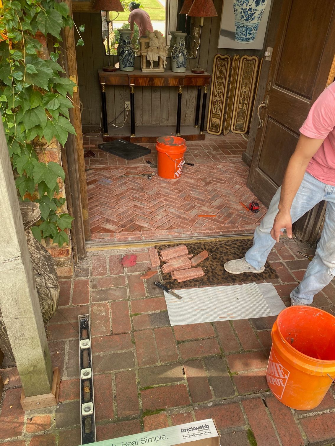 Person laying brick patio; entranceway with vine, orange bucket, level.