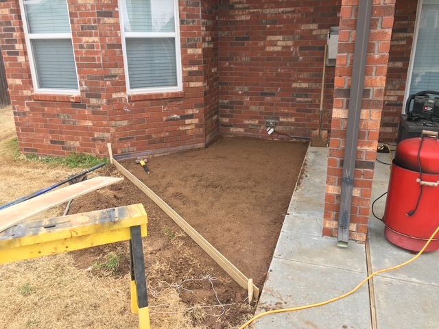 A small patio under construction, with wooden framing, on dirt next to a brick house.