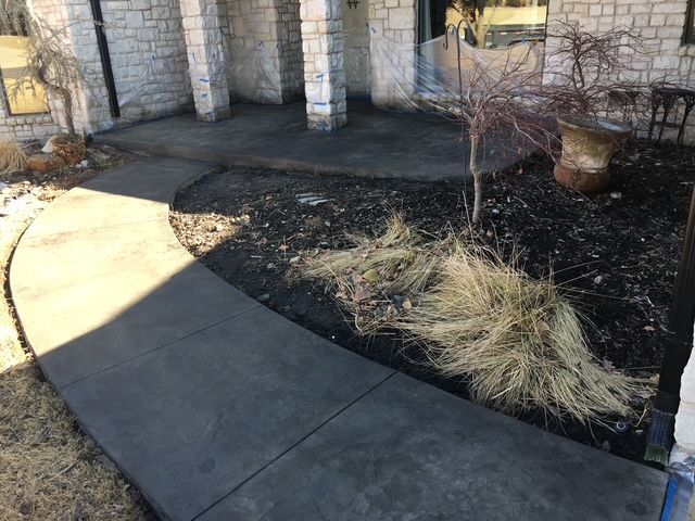 A concrete sidewalk curves toward the front entrance of a building with a small, dry garden.