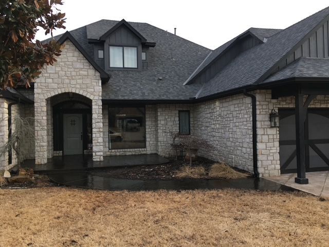 Stone-faced house with dark gray roof, black trim, and a covered porch. Overcast day.