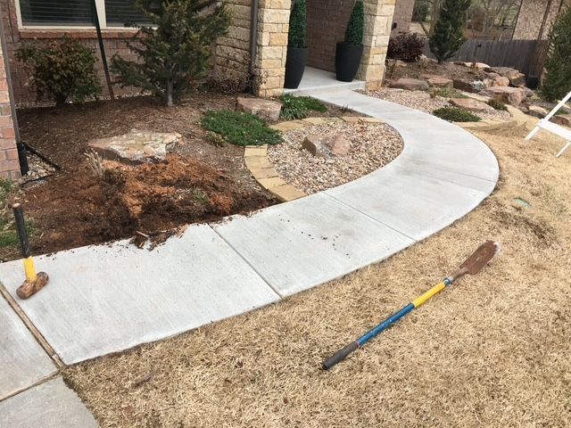 Newly poured concrete walkway curving through a yard with landscaping and a shovel.