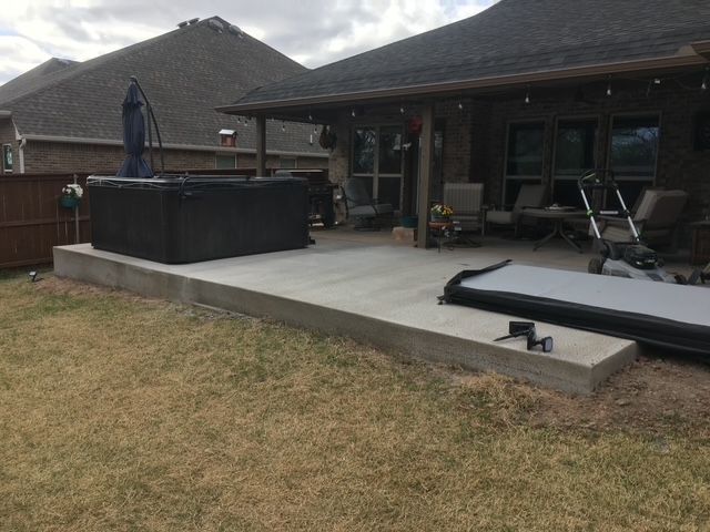 A concrete patio with a hot tub and cover in a backyard setting with a house and brown fence.