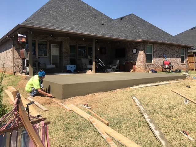 A worker finishes a concrete patio pour next to a house. Green grass, wooden boards, and sunny day.
