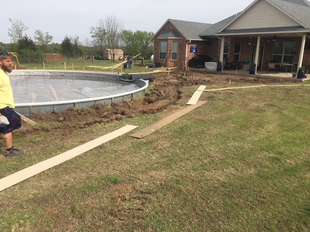 Construction of an in-ground pool next to a house; workers are digging, grass is brown.