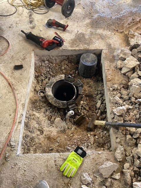 Construction site: Hole in concrete floor exposing a metal pipe with tools and debris. Yellow gloves visible.