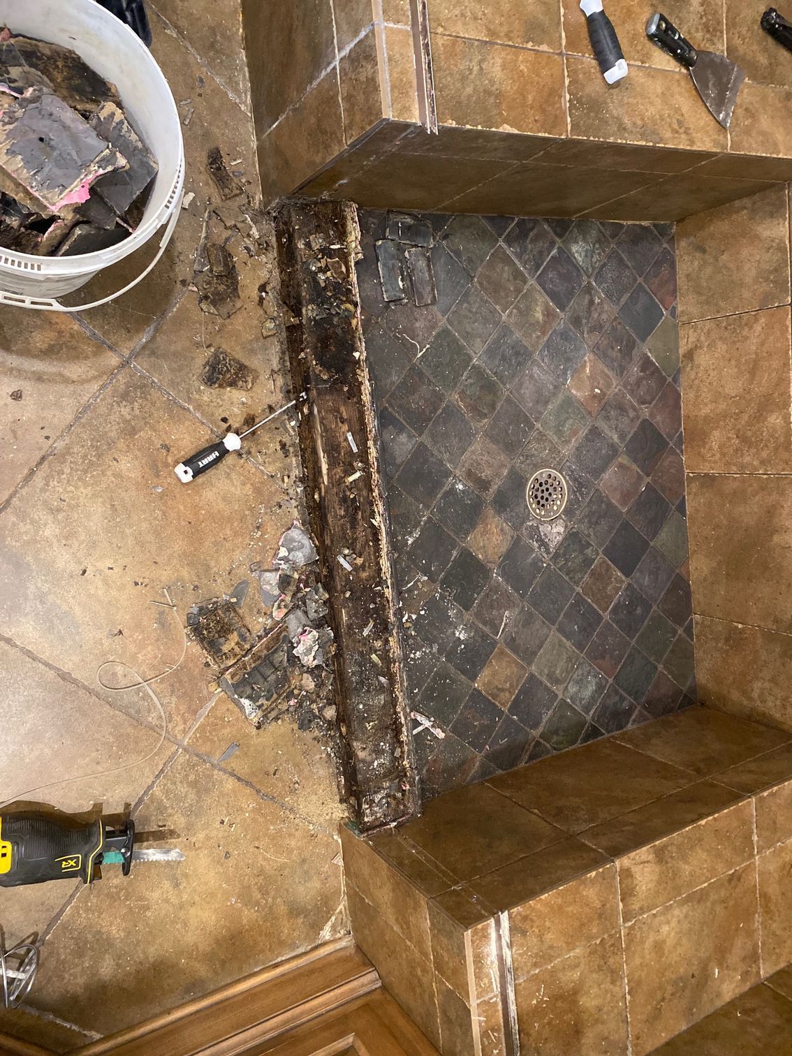 Bathroom shower floor under renovation; damaged tile, wood, and tools.