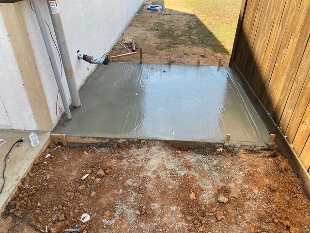 Freshly poured concrete patio next to a building and wooden fence; construction scene.