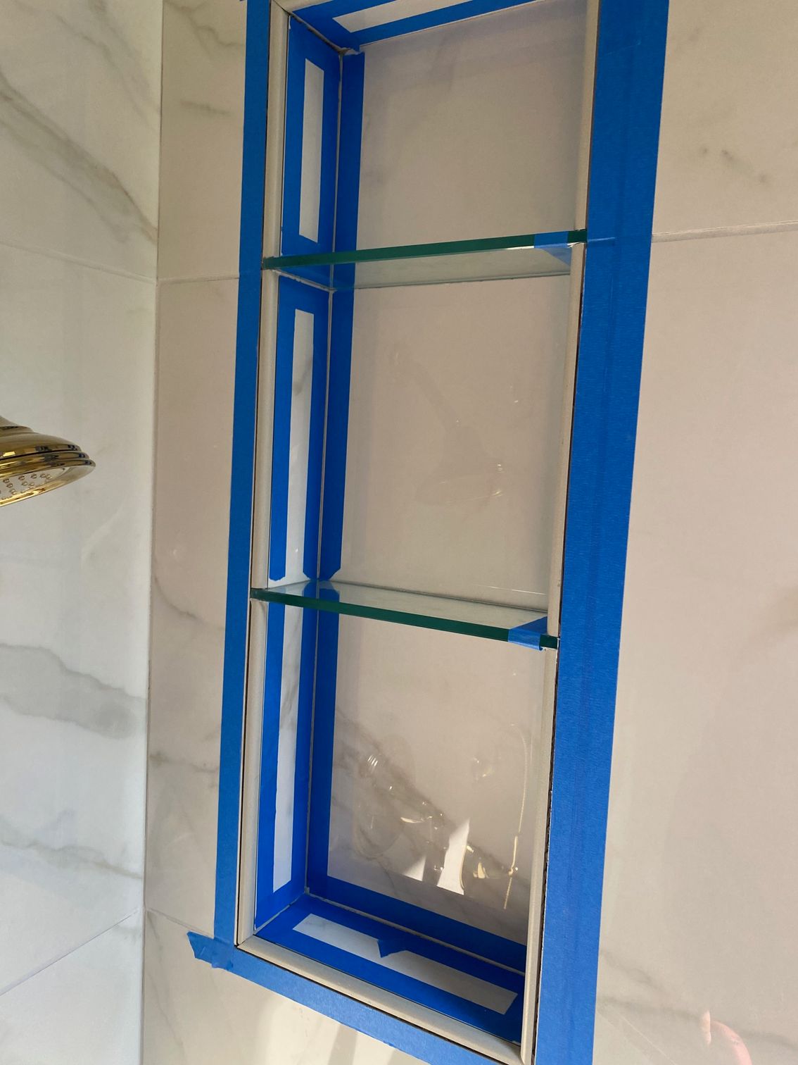 A tiled shower niche with glass shelves, taped with blue painter's tape, awaiting sealant.