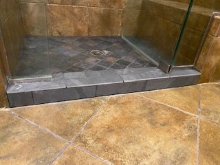 Shower stall with glass door and dark gray tile base and floor. Brown tile flooring surrounds.