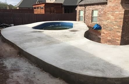 Concrete patio surrounds a pool, next to a brick house and wooden fence.