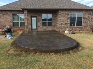 Brown concrete patio outside a brick house. Grass surrounds the patio, a door and windows visible.
