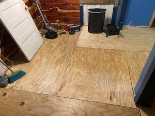 Plywood flooring installed in a room, with a door, tools, and trash can present.