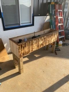 Wooden planter box on legs; exterior setting with ladder and window in background.