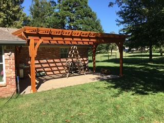 Wooden pergola attached to a brick house, casting shadows on a concrete patio.