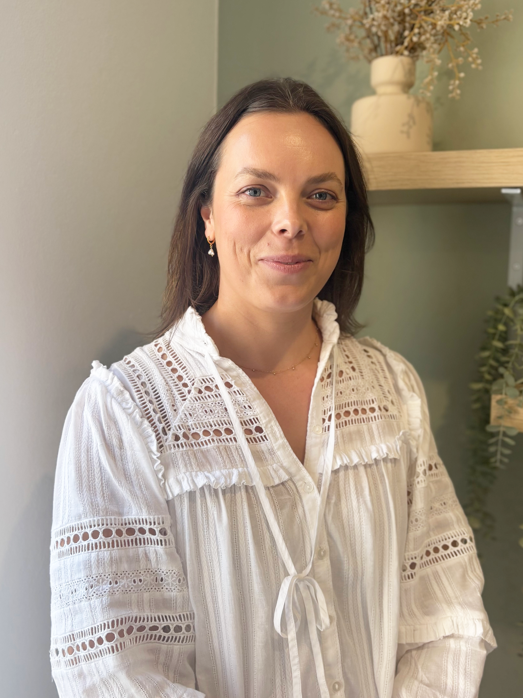 A woman in a white lace shirt is standing in front of a shelf.
