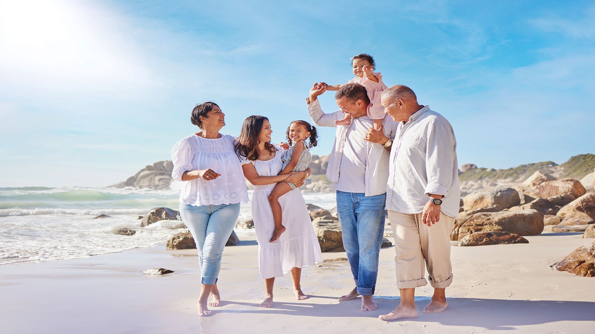 Four people walking on a sunny beach, smiling and talking near the surf.